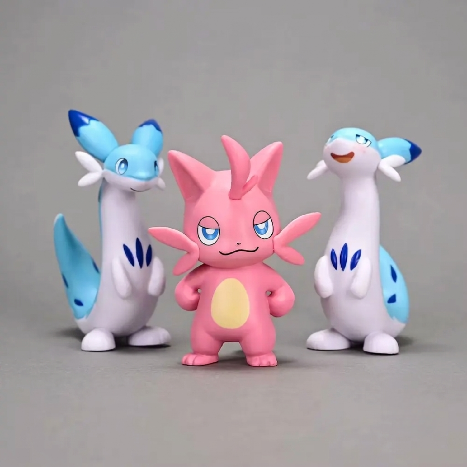 Three Palworld figurines on a gray background: a pink Cattiva stands in the center, flanked by two tall blue-and-white Chillets.