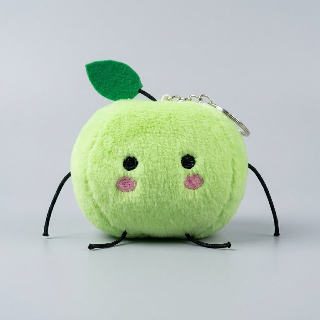A cute, round plush keychain shaped like a smiling green junimo. It features a bright lime green fuzzy body, a small green felt leaf on top, embroidered black eyes and legs, pink cheek patches, and a silver metal keyring attached at the top.