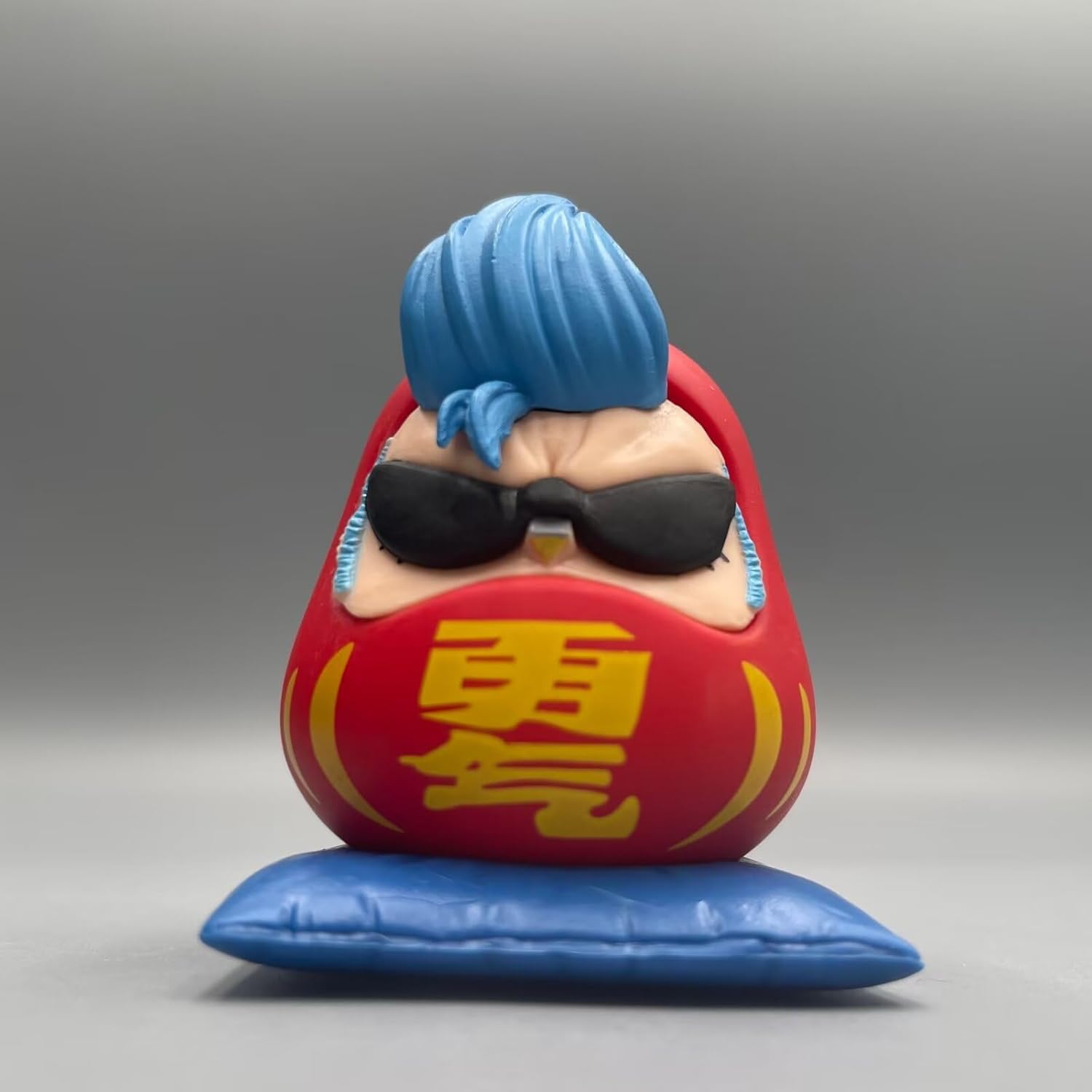 One Piece Daruma figurine of Franky, featuring blue hair, black sunglasses, and a red Daruma body on a blue cushion.