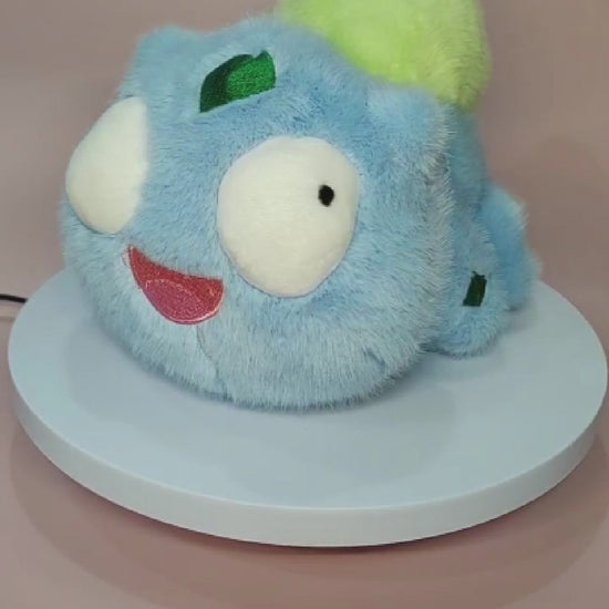 Alt text: Silly "Pokederp" Bulbasaur plushie with a fuzzy light blue body, large round eyes looking in different directions, and a goofy open-mouthed smile. The plush features a soft, bright green bulb on its back and small green patches on its head and body. It sits on a rotating display stand against a pastel pink background.