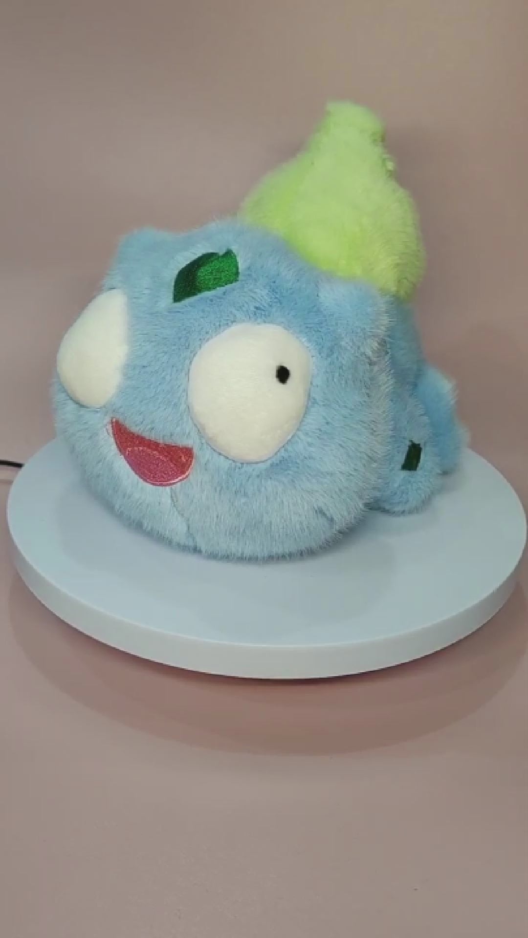 Alt text: Silly "Pokederp" Bulbasaur plushie with a fuzzy light blue body, large round eyes looking in different directions, and a goofy open-mouthed smile. The plush features a soft, bright green bulb on its back and small green patches on its head and body. It sits on a rotating display stand against a pastel pink background.