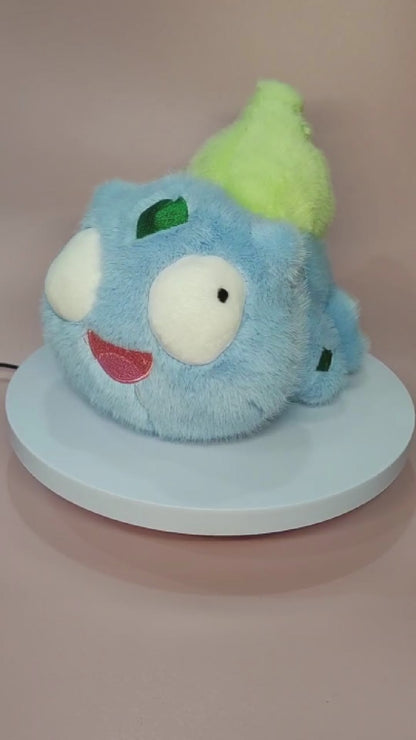 Alt text: Silly "Pokederp" Bulbasaur plushie with a fuzzy light blue body, large round eyes looking in different directions, and a goofy open-mouthed smile. The plush features a soft, bright green bulb on its back and small green patches on its head and body. It sits on a rotating display stand against a pastel pink background.