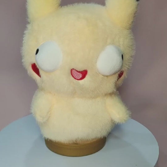 Alt text: Silly "Pokederp" Pikachu plushie with a fuzzy yellow body, oversized round eyes looking in different directions, and a tiny, goofy open-mouthed smile. The plush has black-tipped ears, red cheek patches, and stubby arms. It sits on a rotating display stand against a pastel pink background.