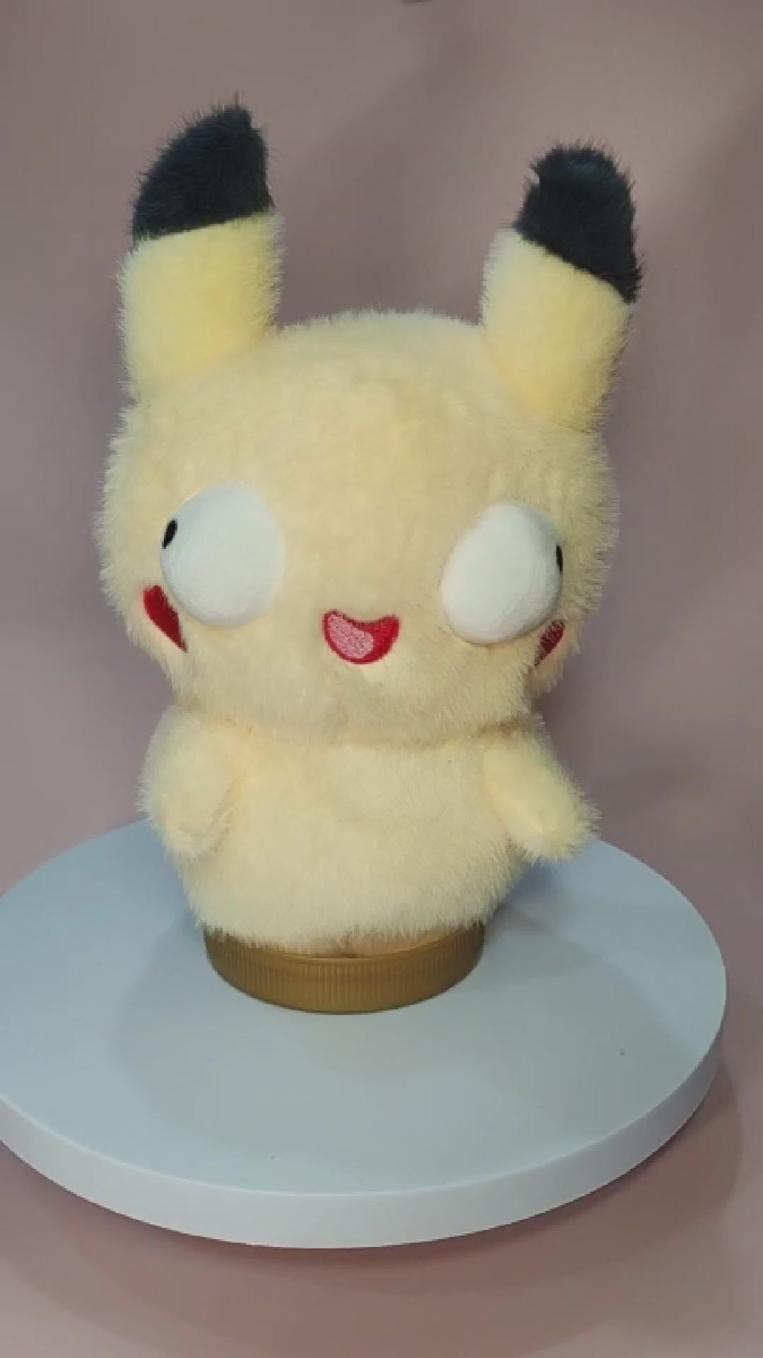 Alt text: Silly "Pokederp" Pikachu plushie with a fuzzy yellow body, oversized round eyes looking in different directions, and a tiny, goofy open-mouthed smile. The plush has black-tipped ears, red cheek patches, and stubby arms. It sits on a rotating display stand against a pastel pink background.