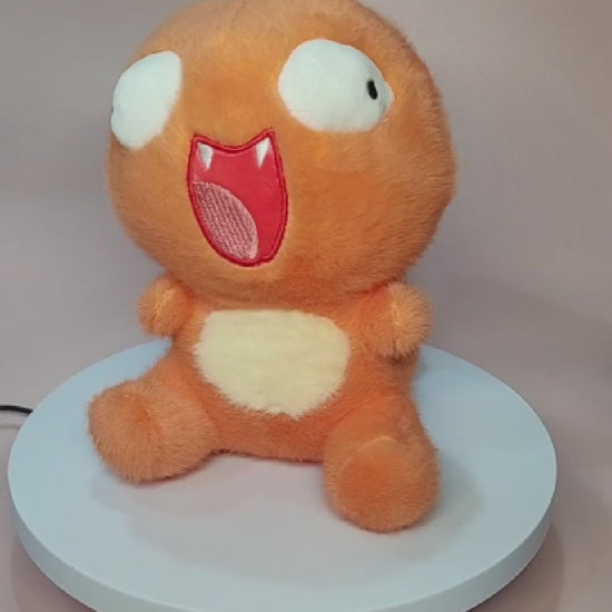 Alt text: Silly "Pokederp" Charmander plushie with a goofy, exaggerated expression, featuring large, uneven eyes and a wide open-mouthed smile. The plush has a soft, fuzzy orange body with a cream-colored belly and stubby arms and legs. It sits on a rotating display stand against a pastel pink background.