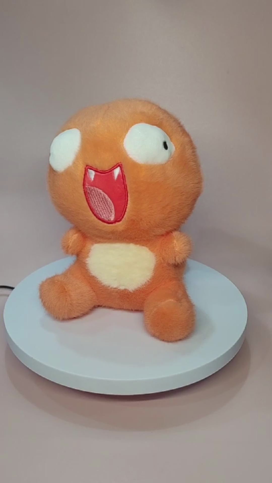 Alt text: Silly "Pokederp" Charmander plushie with a goofy, exaggerated expression, featuring large, uneven eyes and a wide open-mouthed smile. The plush has a soft, fuzzy orange body with a cream-colored belly and stubby arms and legs. It sits on a rotating display stand against a pastel pink background.