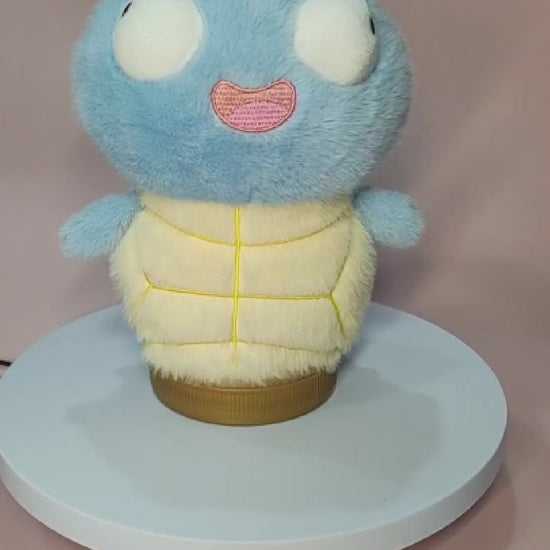 Silly Squirtle plushie with a soft, fuzzy texture, large round eyes, and a goofy open-mouthed expression, sitting on a rotating display stand. The plush features a light blue body, a cream-colored belly with yellow stitching, and small flipper-like arms. The background is a pastel pink surface.