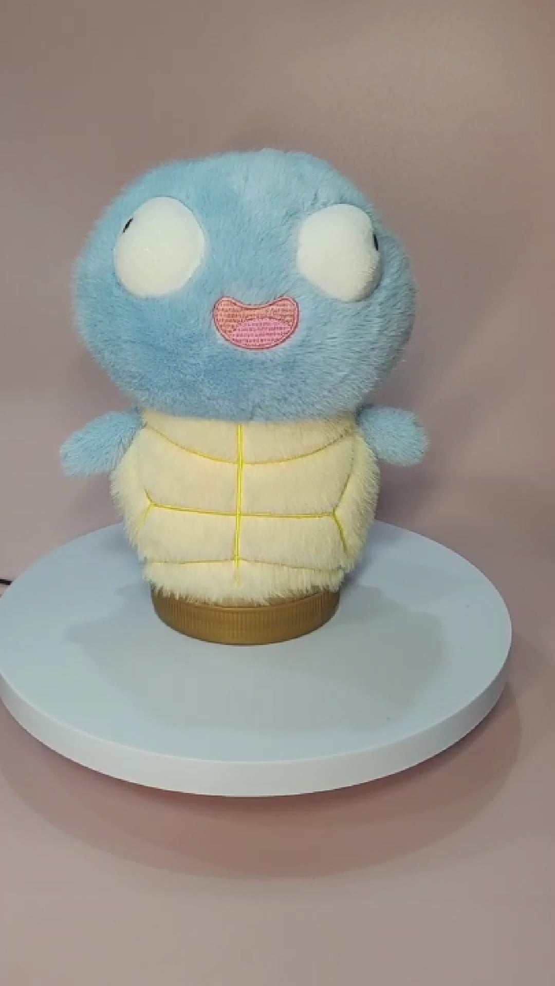 Silly Squirtle plushie with a soft, fuzzy texture, large round eyes, and a goofy open-mouthed expression, sitting on a rotating display stand. The plush features a light blue body, a cream-colored belly with yellow stitching, and small flipper-like arms. The background is a pastel pink surface.