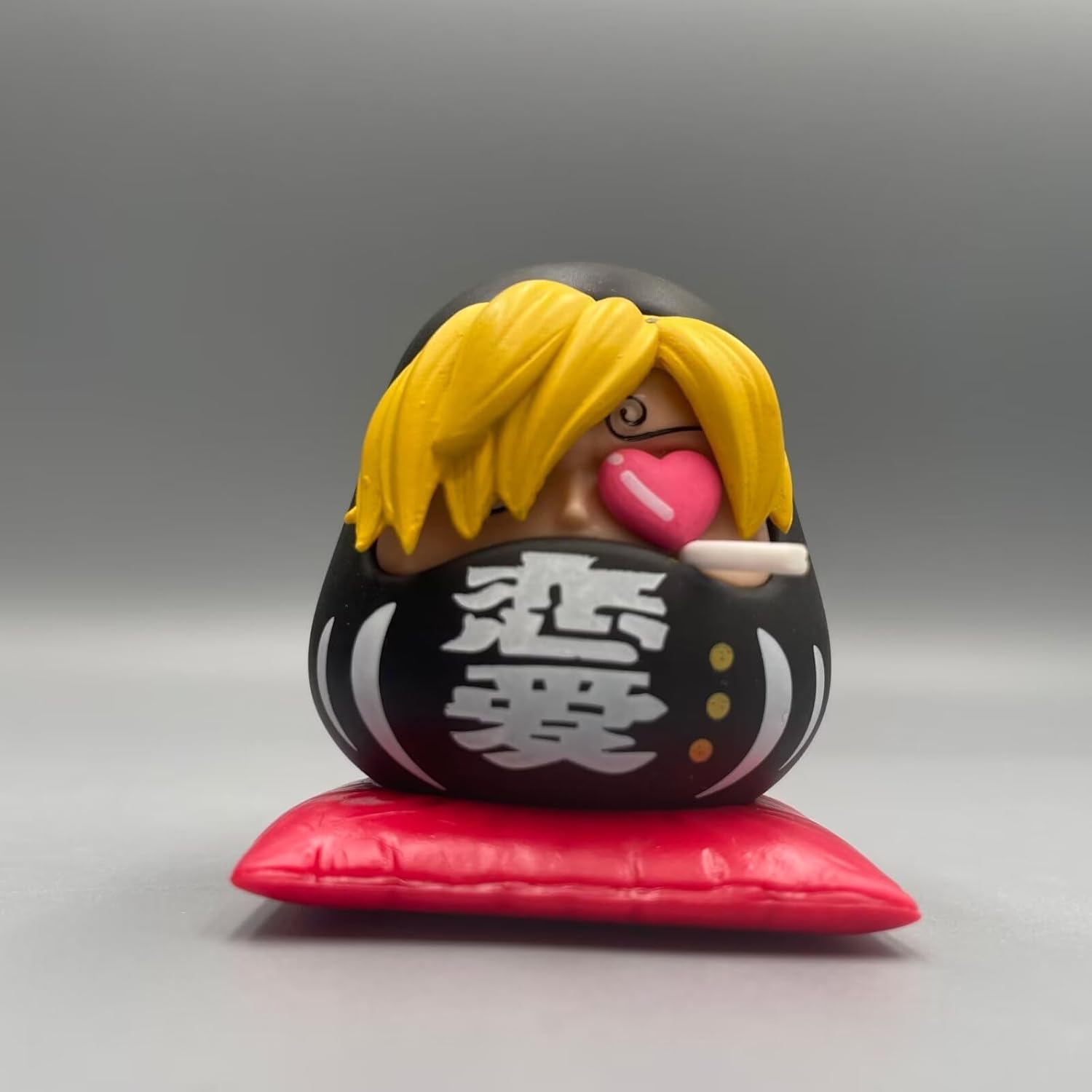 One Piece Daruma figurine of Sanji, featuring blond hair, a heart-shaped mouth, a cigarette, and a black Daruma body on a red cushion.