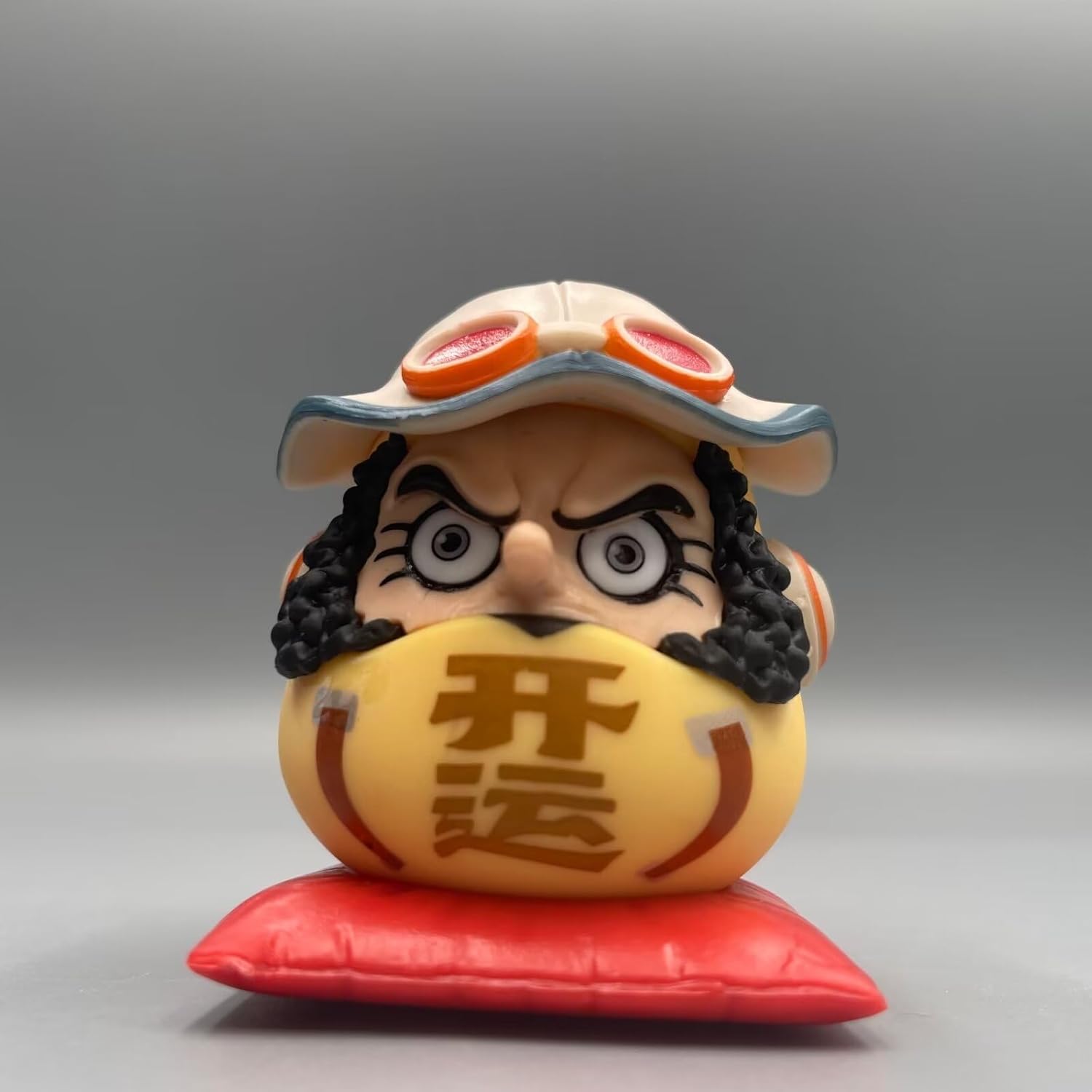 One Piece Daruma figurine of Usopp, featuring curly hair, wide eyes, a pilot hat with goggles, and a yellow Daruma body on a red cushion.