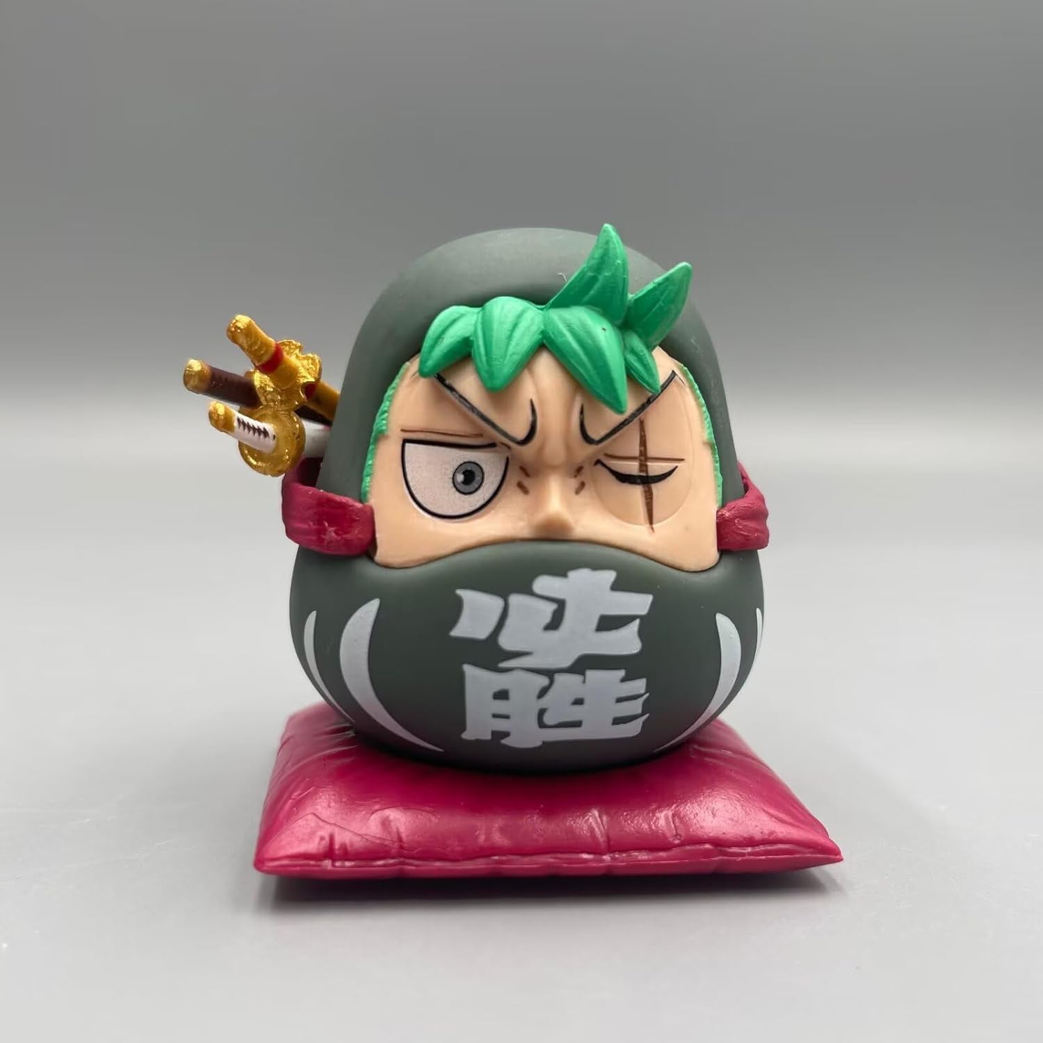 One Piece Daruma figurine of Zoro, featuring green hair, a scarred eye, swords on his back, and a dark Daruma body on a red cushion.